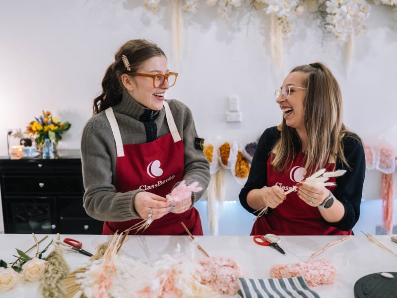 Two women smiling wearing aprons holding flowers at a floristry class.